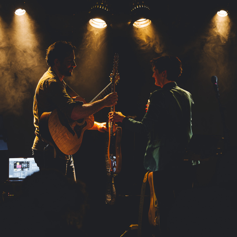 Tamara et Olivier, échangent leurs guitares sur scène sous une lumière dorée et une ambiance enfumée, Ol’d Tam, duo Jukebox