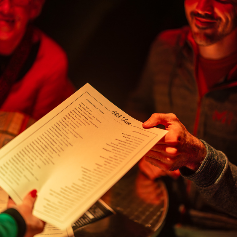 Une main tend un menu de chansons du duo Ol’d Tam à un spectateur, lors d’un concert interactif