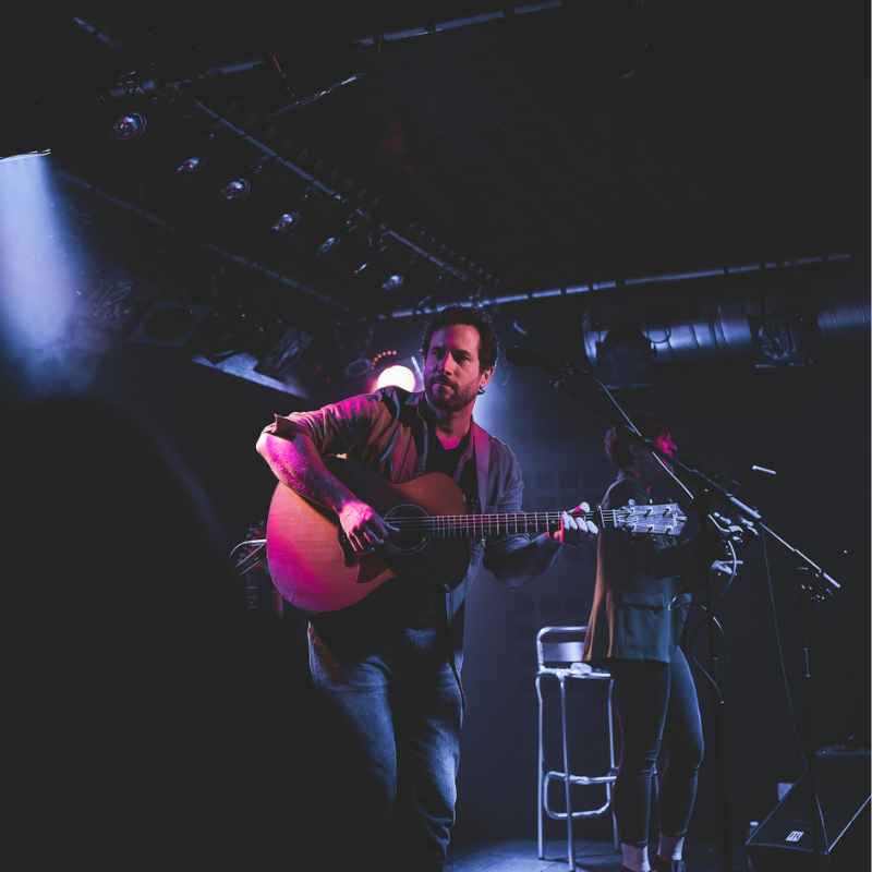 Olivier, guitariste du duo Ol’d Tam, joue de la guitare acoustique sur scène, Tamara en arrière-plan à la basse