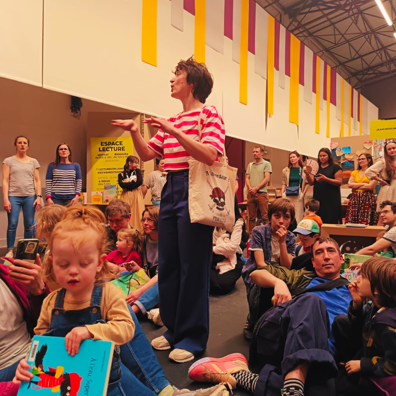 La cheffe de chœur de la Chorale Pirate s’adresse à des parents et enfants lors de la Fête du Livre Jeunesse de Villeurbanne, entourée de choristes et d’un jeune public assis.