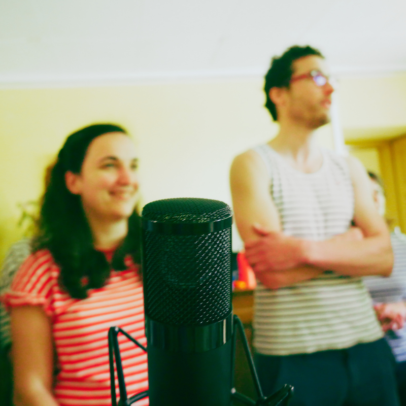 Un micro de studio au premier plan, avec deux choristes de la Chorale Pirate souriants en arrière-plan.