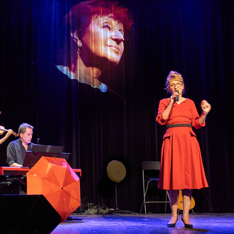 Isabelle Desmero interprète une chanson du spectacle Chansons & Coquelicots, hommage à Anne Sylvestre, accompagnée par Alexandre Afonso au piano, avec le visage d’Anne Sylvestre projeté en arrière-plan.