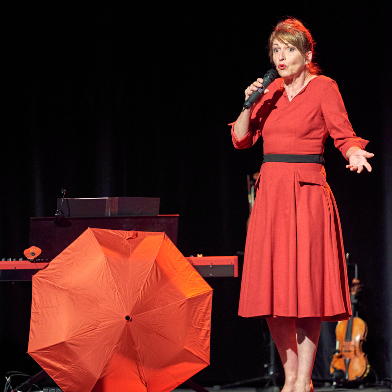 Isabelle Desmero interprète une chanson sur scène lors du spectacle "Chansons & Coquelicots", hommage à Anne Sylvestre, avec un parapluie rouge ouvert au pied du piano.