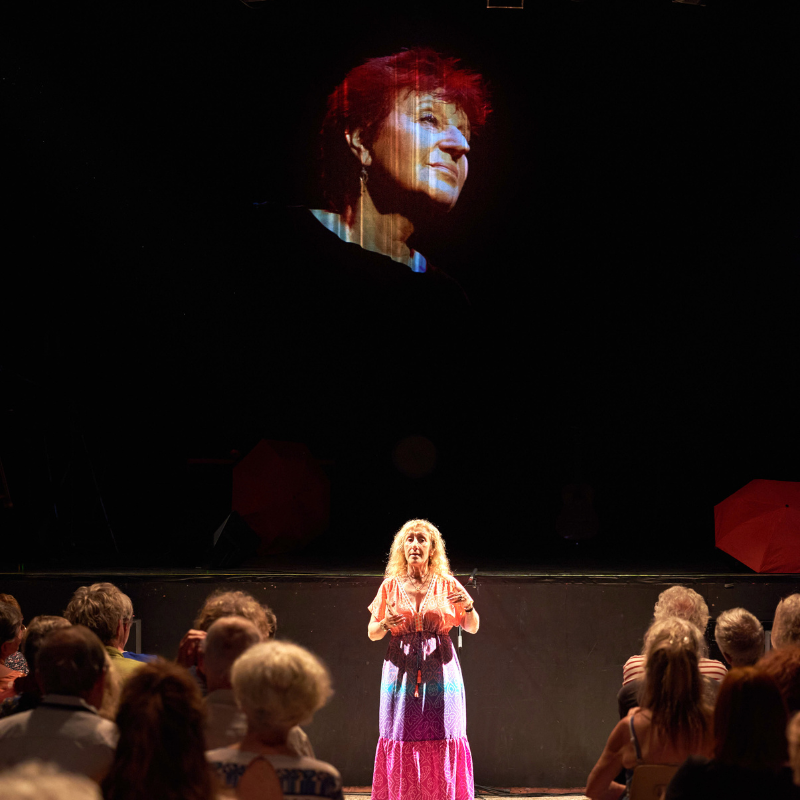 La programmatrice culturelle de Gardanne prend la parole avant la première du spectacle Chansons & Coquelicots, avec en arrière-plan la photo d’Anne Sylvestre projetée sur un rideau noir.
