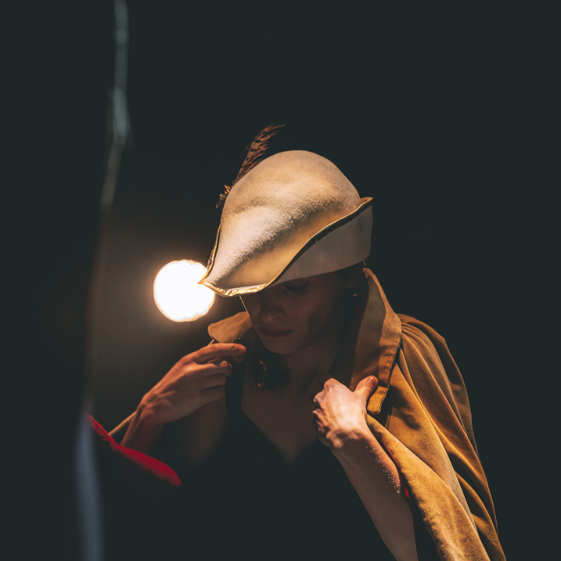 Interprète en chapeau et cape, se préparant sous la lumière d’un projecteur dans Splendeur & Décadence