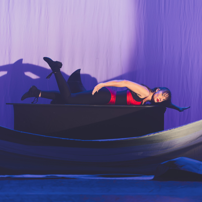 Interprète allongée sur une table, ombre de requin projetée derrière elle dans Splendeur & Décadence