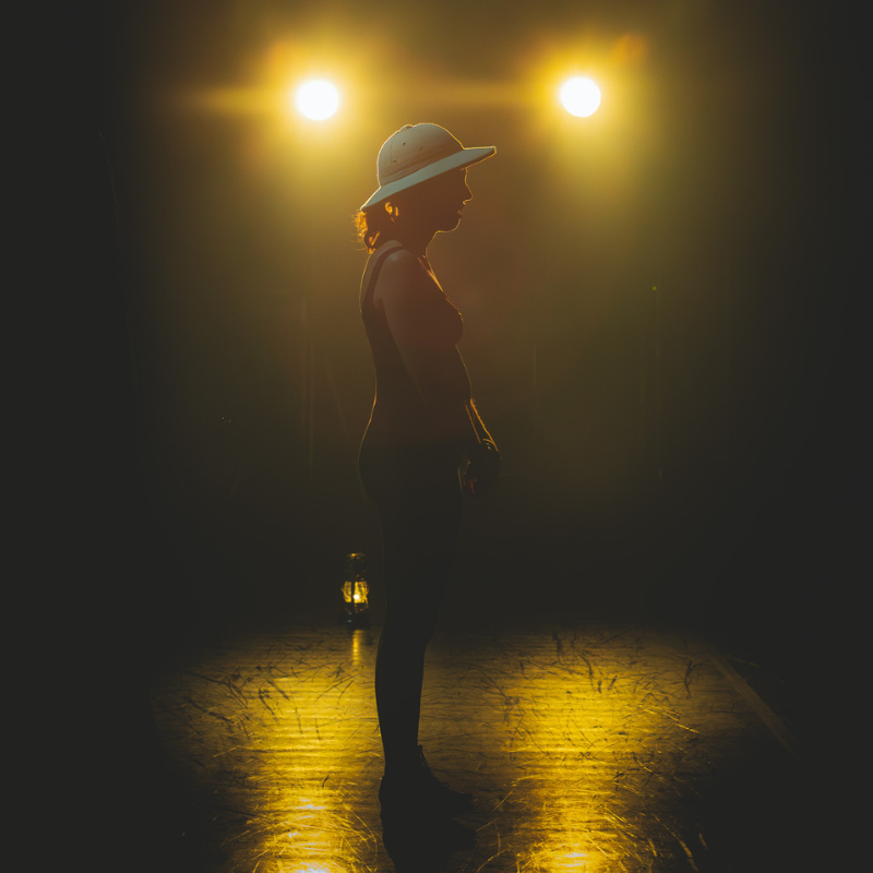 Silhouette d’une interprète coiffée d’un casque colonial, en contre-jour sur scène dans Splendeur & Décadence