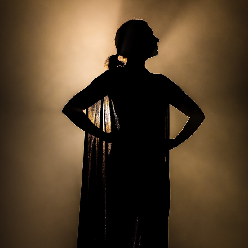 Silhouette d’une interprète en cape, mains sur les hanches, éclairée par un halo doré dans Splendeur & Décadence