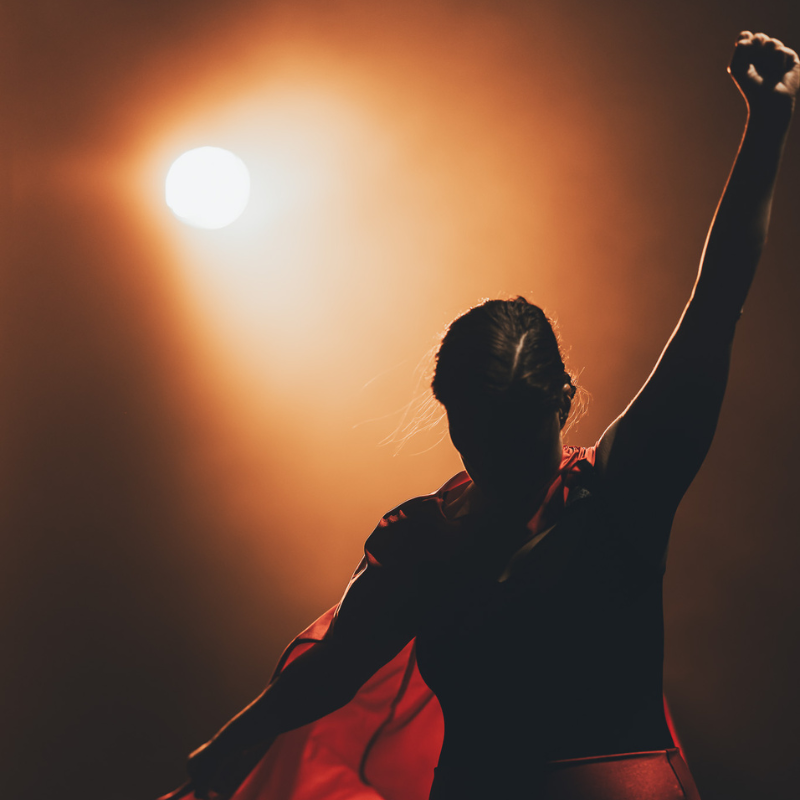 Silhouette d’une interprète en cape rouge, poing levé, éclairée par un projecteur dans le spectacle Splendeur & Décadence