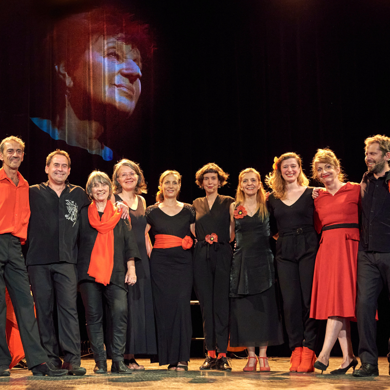 L’ensemble des artistes et techniciens du spectacle Chansons & Coquelicots, hommage à Anne Sylvestre, posent souriants sur scène avec en arrière-plan le visage d’Anne Sylvestre projeté.