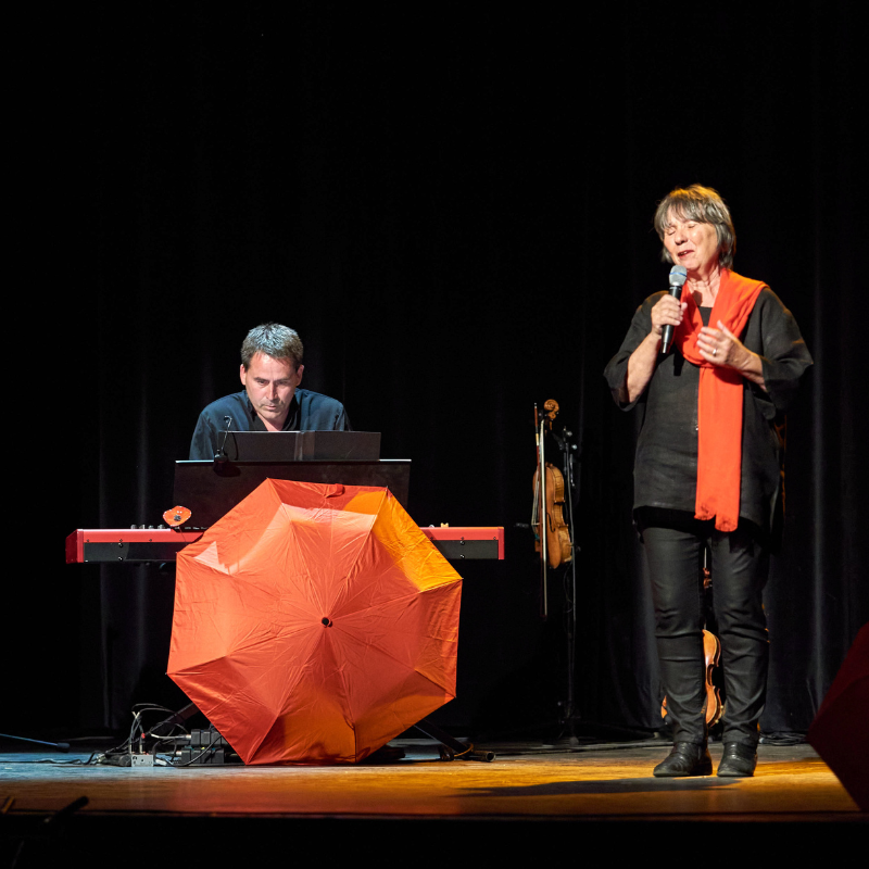 Martine Scozzesi chante sur scène lors du spectacle "Chansons & Coquelicots", hommage à Anne Sylvestre, accompagnée d'Alexandre Afonso au piano, avec un parapluie rouge au pied du piano.